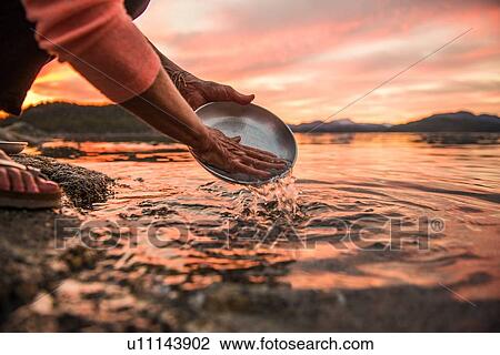 Stock Image - Mature woman washing plate at sunset, cropped, Quadra Island, Campbell River, Canada. Fotosearch