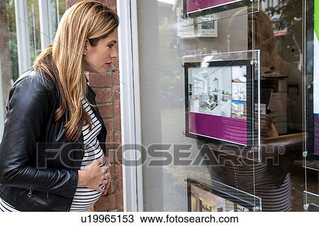Stock Image - Pregnant mid adult woman looking through estate agents window. Fotosearch