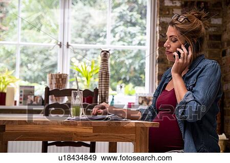 Stock Photo - Pregnant mid adult woman making smartphone call in kitchen. Fotosearch