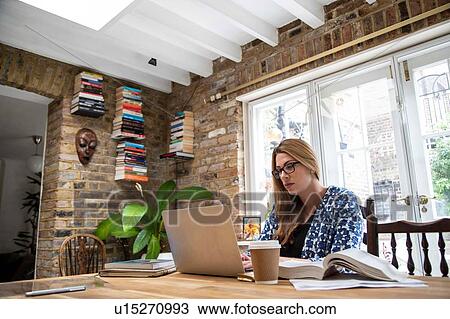 Stock Image - Pregnant mid adult woman typing on laptop at kitchen table. Fotosearch