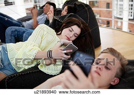 Stock Image - School friends lying on beanbags and texting at school. Fotosearch