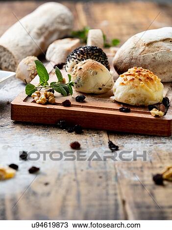 Stock Image - Seeded bread rolls and rustic loaves on chopping board, still life. Fotosearch