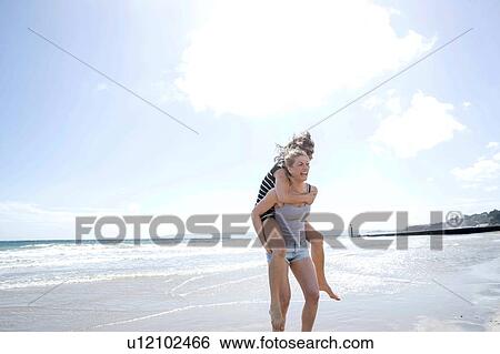 Stock Photograph - Sisters playing piggyback on beach. Fotosearch