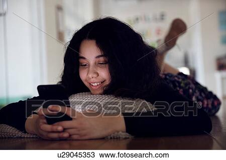 Stock Image - Teenage girl lying on cushion on floor reading text message. Fotosearch