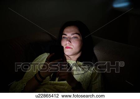 Stock Image - Teenage girl lying on sofa in dark reading text message. Fotosearch