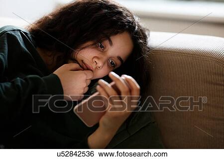 Stock Photograph - Teenage girl lying on sofa in dark reading text message. Fotosearch