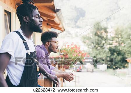 Stock Image - Two cool young men looking out from log cabin porch, Primaluna, Trentino-Alto Adige, Italy. Fotosearch