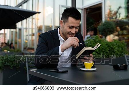 Stock Image - Young businessman at sidewalk cafe looking at notebook, Milan, Italy. Fotosearch