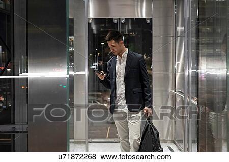 Stock Photo - Young businessman in office building at night looking at smartphone, Milan, Italy. Fotosearch