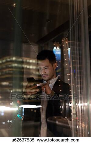 Stock Photography - Young businessman in office building at night looking at smartphone, window view, Milan, Italy. Fotosearch