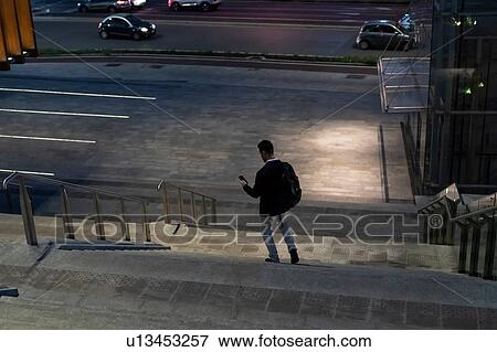 Stock Photo - Young businessman moving down city stairway at night looking at smartphone, Milan, Italy. Fotosearch