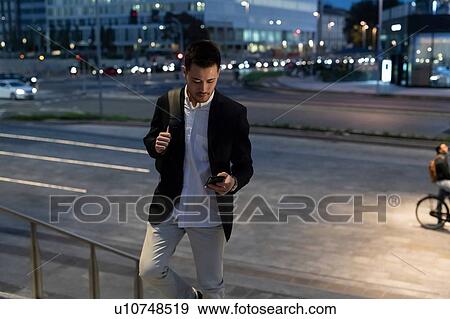 Stock Photo - Young businessman moving up city stairway looking at smartphone, Milan, Italy. Fotosearch