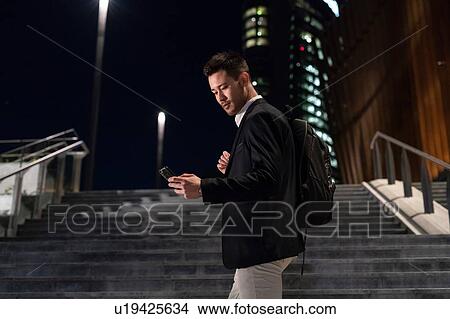 Picture - Young businessman on city stairway at night looking at smartphone, Milan, Italy. Fotosearch