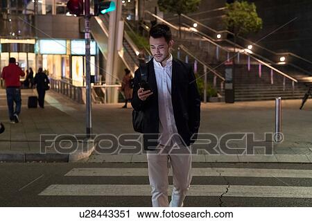 Stock Image - Young businessman on pedestrian crossing at night looking at smartphone, Milan, Italy. Fotosearch
