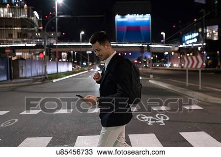 Stock Image - Young businessman on pedestrian crossing at night looking at smartphone, Milan, Italy. Fotosearch