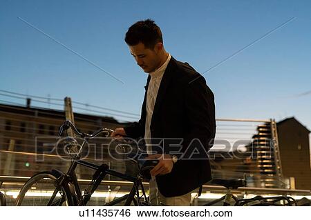 Stock Photograph - Young businessman picking up city bike at night, Milan, Italy. Fotosearch