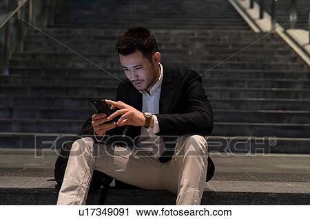Stock Image - Young businessman sitting on city stairway at night looking at smartphone, Milan, Italy. Fotosearch