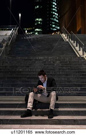 Stock Photography - Young businessman sitting on city stairway at night using smartphone touchscreen, Milan, Italy. Fotosearch