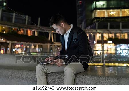Stock Photograph - Young businessman sitting on city wall at night using laptop, Milan, Italy. Fotosearch
