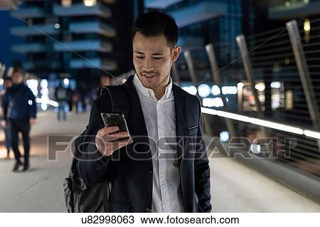 Stock Image - Young businessman walking on city street at night looking at smartphone, Milan, Italy. Fotosearch