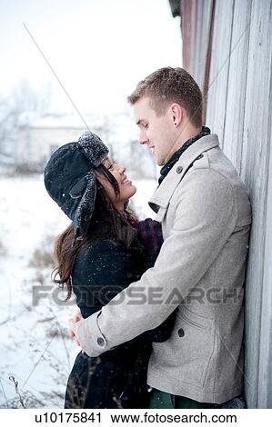 Stock Image - Young couple hugging against cabin in snow. Fotosearch