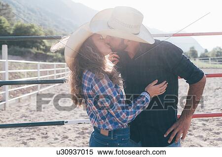 Stock Photography - Young cowgirl and boyfriend kissing in rural equestrian arena. Fotosearch