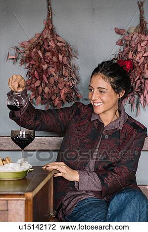 Stock Image - Young woman at rustic table pouring red wine. Fotosearch