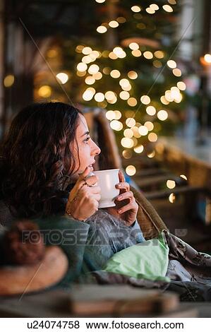 Stock Photo - Young woman drinking coffee in cafe with decorative lights. Fotosearch
