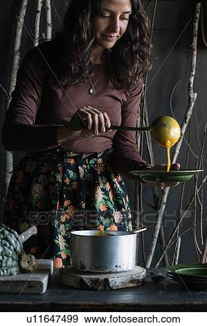 Stock Photo - Young woman ladling fresh soup from saucepan at rustic kitchen counter. Fotosearch