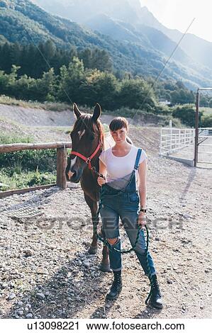 Stock Image - Young woman leading horse in rural equestrian arena, full length portrait. Fotosearch