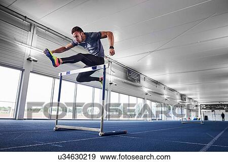 Athlete jumping over hurdle on indoor running track View Large Photo Image Stock Photo - Athlete jumping over hurdle on indoor running track. Fotosearch