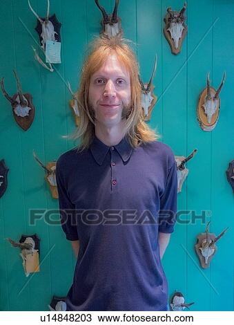Stock Image - Barber posing against green wall mounted with animal skulls. Fotosearch