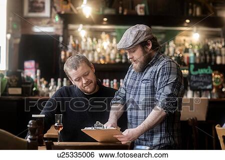 Stock Photograph - Barman looking at menu with customer in traditional Irish public house. Fotosearch