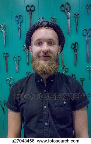 Stock Photograph - Bearded barber posing against green wall mounted with scissors. Fotosearch