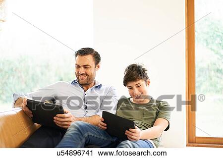 Stock Image - Boy and father looking at digital tablets whilst reclining on sofa. Fotosearch