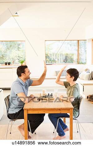 Stock Photograph - Boy and father playing chess at living room table, high fiving. Fotosearch