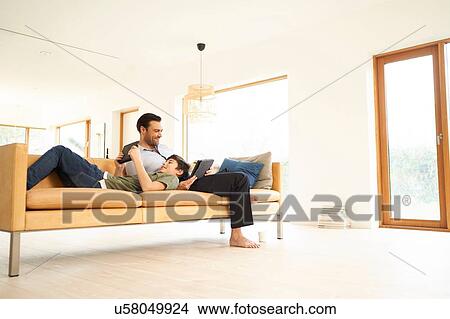 Picture - Boy and father using digital tablets whilst reclining on sofa. Fotosearch