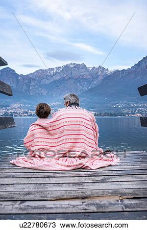 Stock Image - Boy and father wrapped in blanket on lakeside pier, rear view, Lake Como, Onno, Lombardy, Italy. Fotosearch