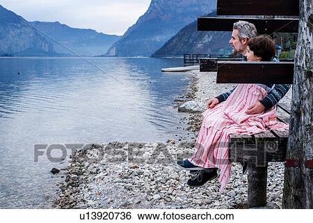 Stock Photograph - Boy and father wrapped in blanket on lakeside pier, side view, Lake Como, Onno, Lombardy, Italy. Fotosearch
