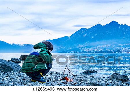 Stock Image - Boy by campfire at dusk, Onno, Lombardy, Italy. Fotosearch