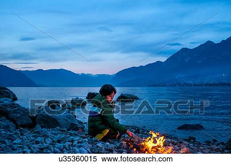 Stock Photography - Boy by campfire at dusk, Onno, Lombardy, Italy. Fotosearch
