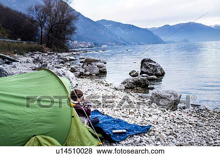 Stock Photo - Boy sitting on blanket by tent drinking coffee on lakeside, Lake Como, Onno, Lombardy, Italy. Fotosearch
