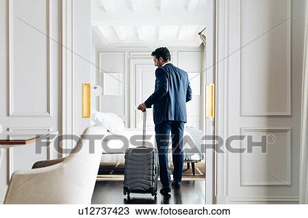 Stock Image - Businessman with wheeled luggage in suite. Fotosearch
