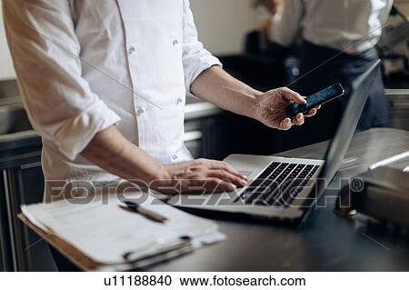 Stock Image - Chef using laptop and cellphone in kitchen. Fotosearch
