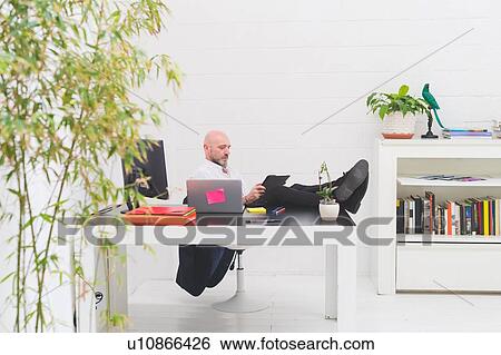 Stock Photograph - Cool businessman with feet up in office looking at digital tablet. Fotosearch