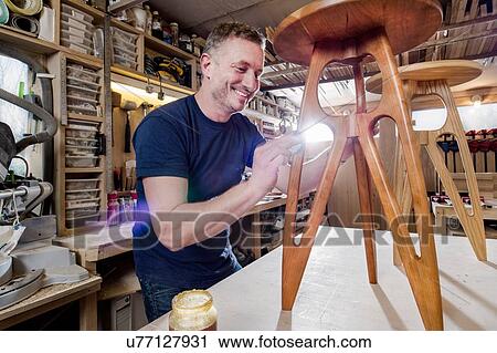 Stock Image - Craftsman polishing wooden stool with oil finish. Fotosearch
