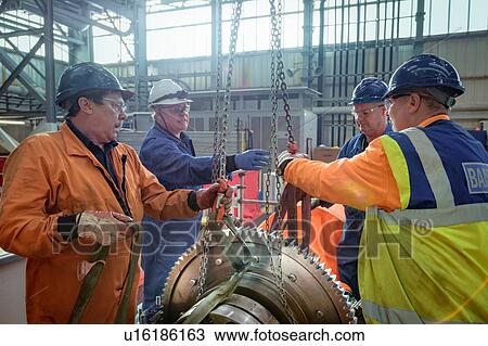 Stock Image - Engineers craning large gear into place in turbine hall of nuclear power station. Fotosearch