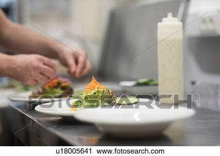 Stock Image - Fast food worker adding salad to hamburger in commercial kitchen, detail of hand. Fotosearch