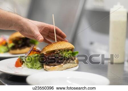 Stock Photograph - Fast food worker preparing hamburger and salad in commercial kitchen, close up of hand. Fotosearch