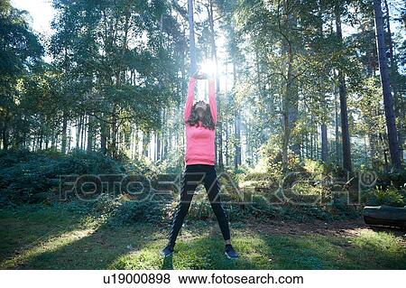 Stock Photo - Female runner stretching arms and warming up in sunlit forest. Fotosearch